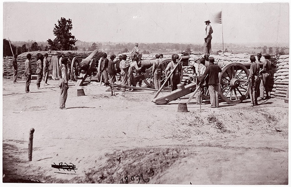 A black-and-white image of men surrounding cannons against a sandbag wall with the American flag planted on the wall