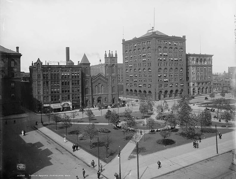 A black-and-white photograph of a public square with a fountain in the middle; across the street, a 10-story building