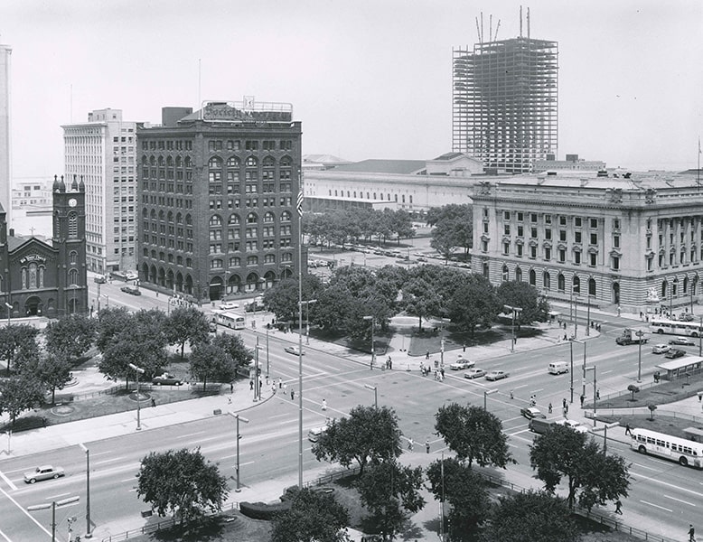 A black-and-white photograph of a city intersection with buildings in the background and a skyscraper in the process of being constructed.