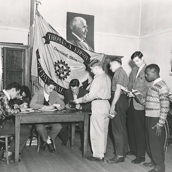 A black-and-white photograph of a line of boys waiting near a desk with a flag behind it in a classroom.