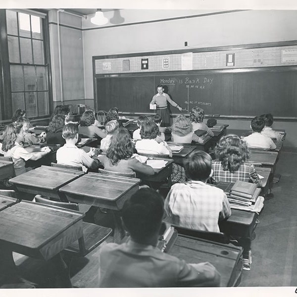 A black-and-white photo of a classroom with children sitting at their desks with a teacher at the chalkboard in front of the classroom.