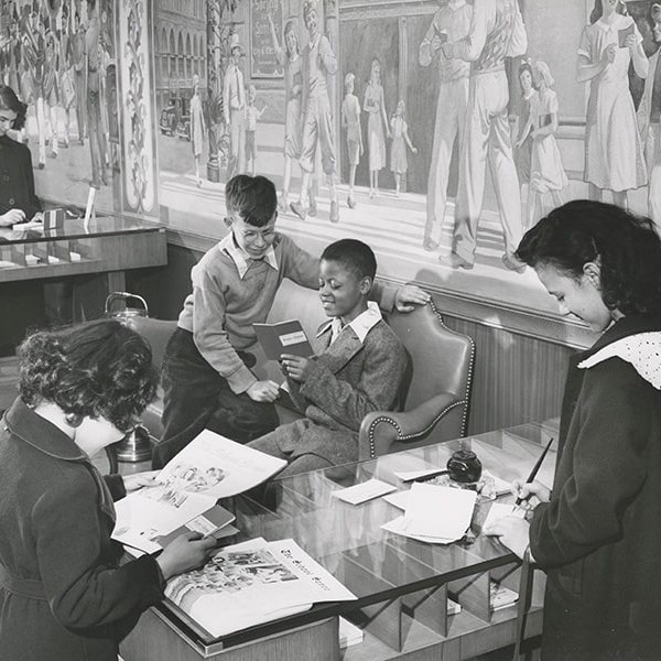 A black-and-white photograph of a classroom setting with two boys on a couch reading books; in the foreground, two girls at a counter reading books.