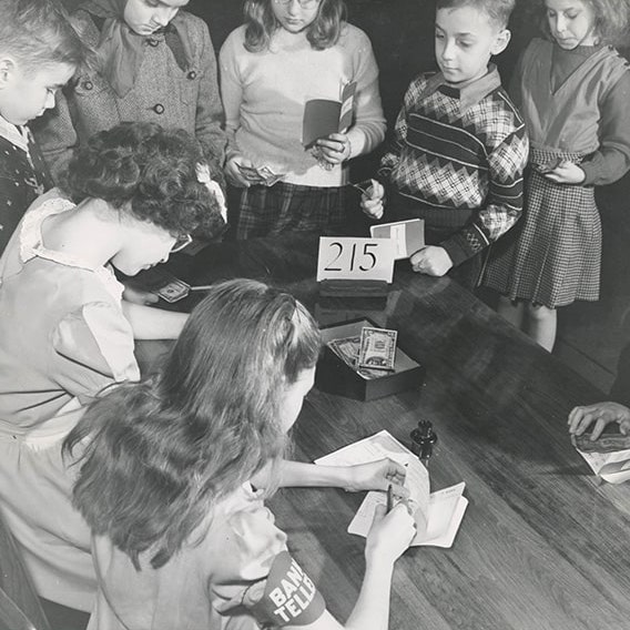 A black-and-white photograph of children sitting and standing around a desk, writing on pieces of paper.