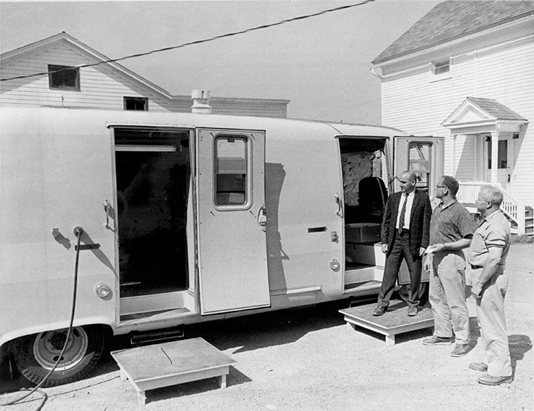 A black-and-white photograph of a mobile banking unit parked in a suburban neighborhood with three men standing at the entrance in a line.