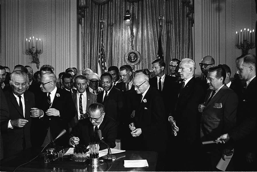 A black-and-white photograph of President Johnson alongside Martin Luther King and others as the president signs the Civil Rights Act in the White House.