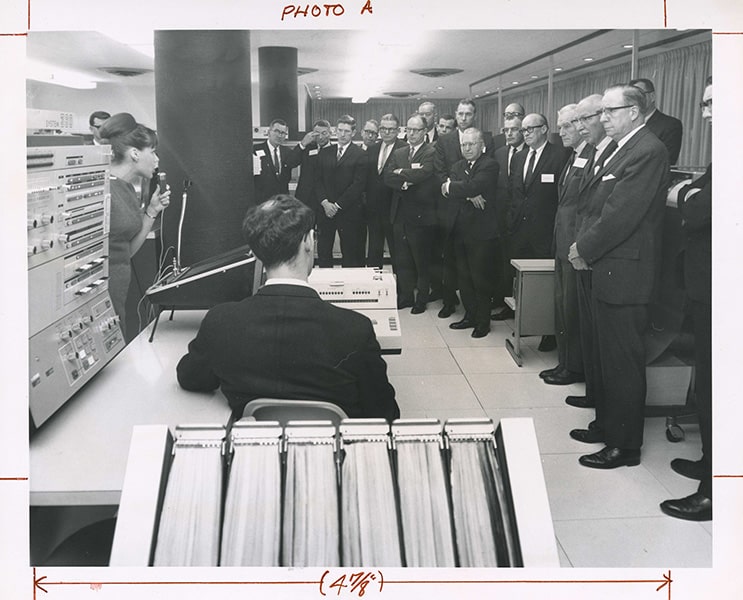 A black-and-white photograph of an assembly of men in suits in a room with a large computer. In the foreground, a man is sitting at a table with a typewriter, and a woman to his left with a microphone.