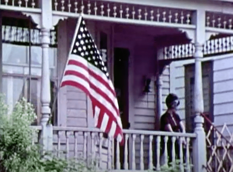 A colored image of the front porch of a home with the American flag in the foreground and a woman sitting on the porch in the background.