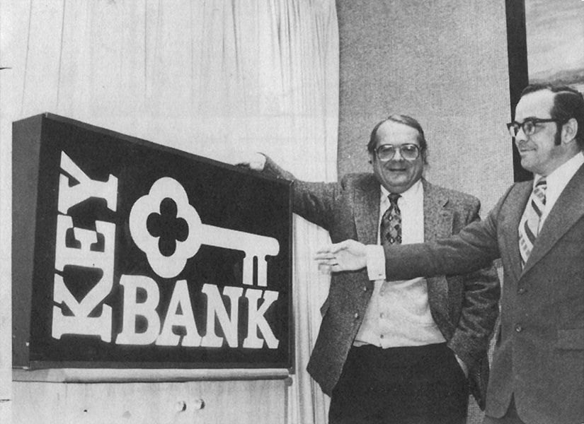 A black-and-white photo of two men standing next to and showcasing a large KeyBank sign.