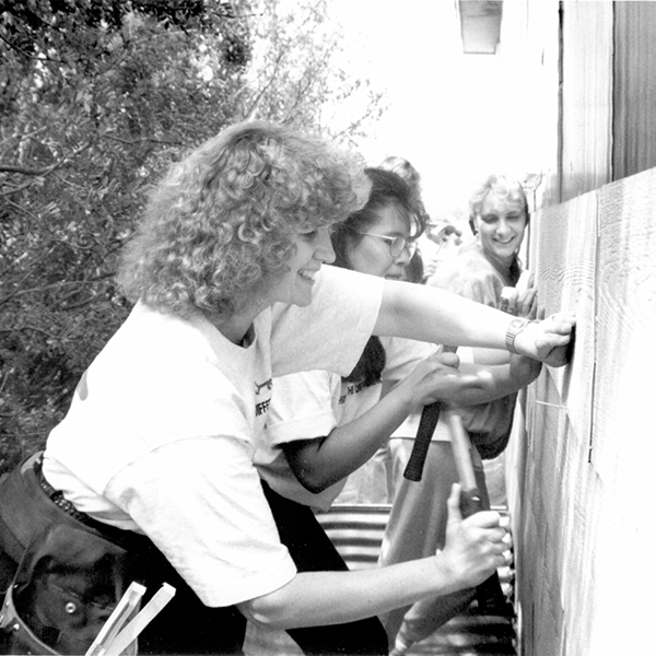 A black-and-white photograph of a volunteering woman hammering in a nail on the side of a house with others behind her.