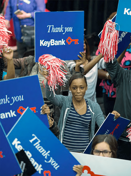 A colored photograph of people in a crowd holding blue signs with 'Thank you, KeyBank.' In focus in the middle is a woman with a pom-pom.